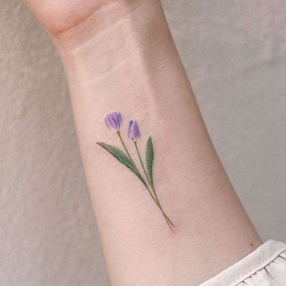 A delicate watercolor tulip tattoo on the wrist, featuring two soft purple flowers with fine green stems and leaves for a natural, elegant look.