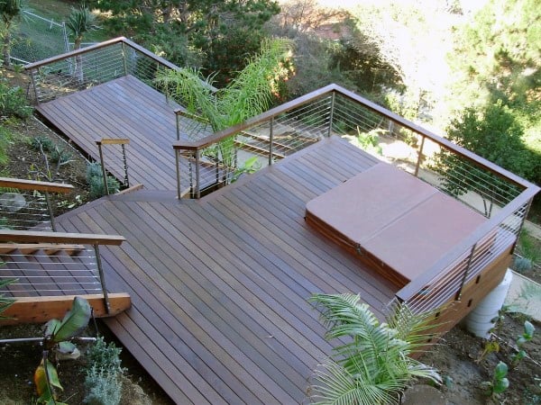 Multi-level wooden deck with cable railing, built-in hot tub, and lush greenery surroundings.