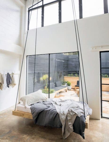 Minimalist hanging bed with black rope supports in a bright, two-story space overlooking a patio.