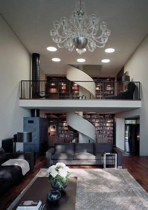 Modern living room with a spiral staircase, chandelier, bookshelves, and dark furniture, leading to an upper-level library