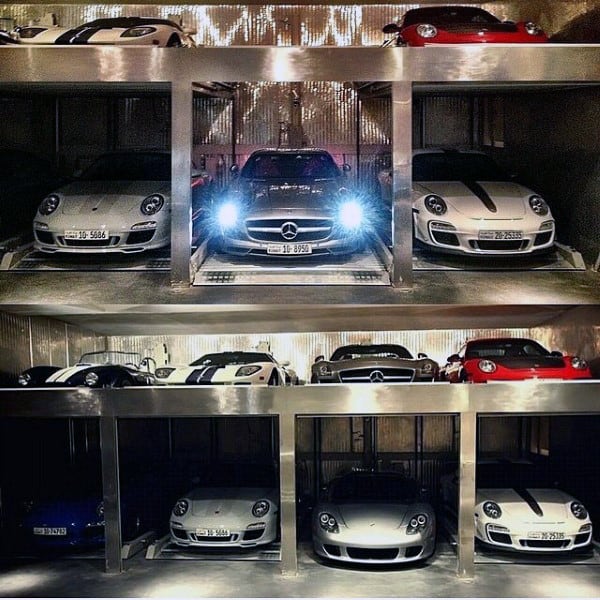 A sleek, two-story dream garage with an organized layout featuring luxury cars, including Porsches and Mercedes, highlighted with dramatic lighting.