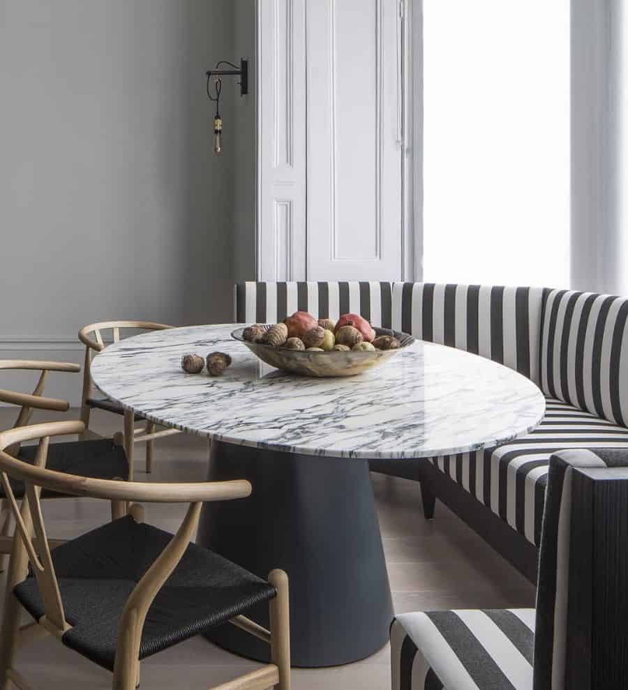 Marble oval dining table with black striped banquette and wooden chairs in modern setting.