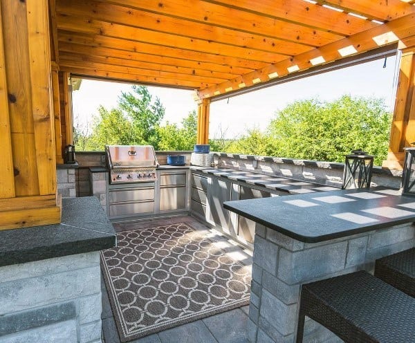 Outdoor kitchen featuring a grill, stone countertops, and a wooden pergola with lush greenery and brickwork in the background