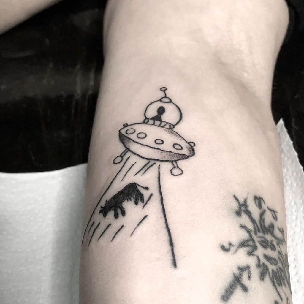 A black ink tattoo of a UFO beaming up a cow, drawn in a simple, sketch-like style on the forearm.