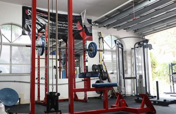 Garage gym with a red power rack, cable machine, weight plates, and suspension trainer setup.