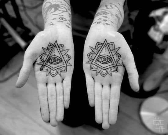 Matching tattoos on both palms featuring an all-seeing eye inside a triangle with a mandala-style border, inked in black.