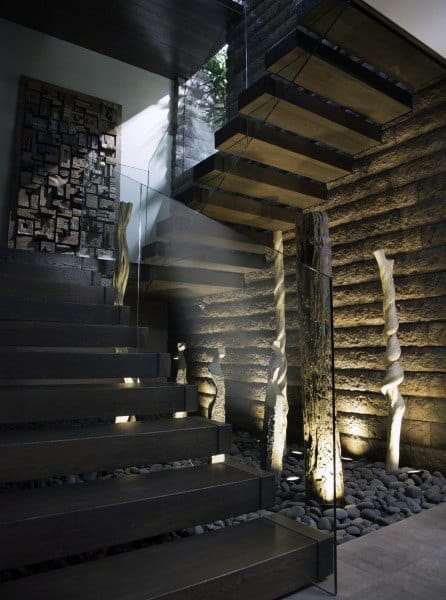 Modern staircase with floating wooden steps, glass railing, and stone wall featuring artistic lighting and sculptures
