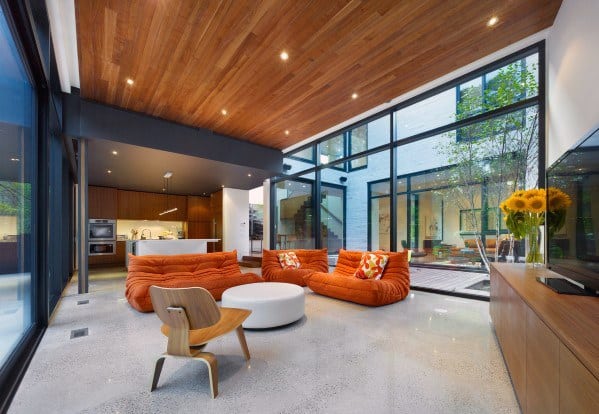 Modern living room with wood ceiling, orange sofas, large windows, and an open kitchen.
