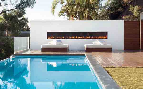 Modern poolside with two loungers and a long, narrow fireplace on a patio surrounded by greenery