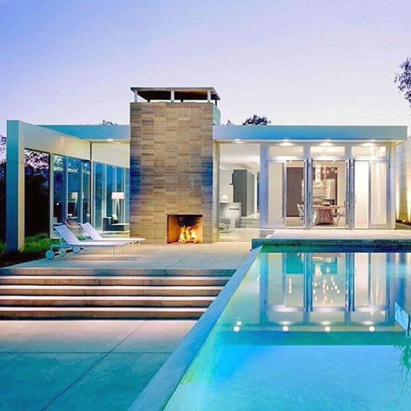 Modern glass house with a lit fireplace, patio, and pool at sunset