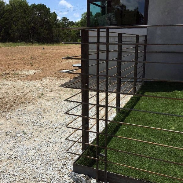 Modern outdoor space with a metal railing, fence, artificial grass, bordered by gravel and a dirt area