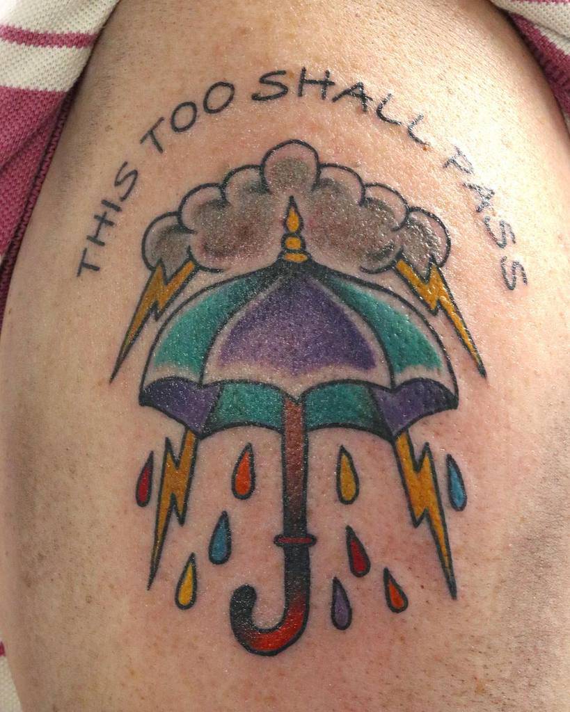 Tattoo with "THIS TOO SHALL PASS" and an umbrella, surrounded by lightning and colorful raindrops.