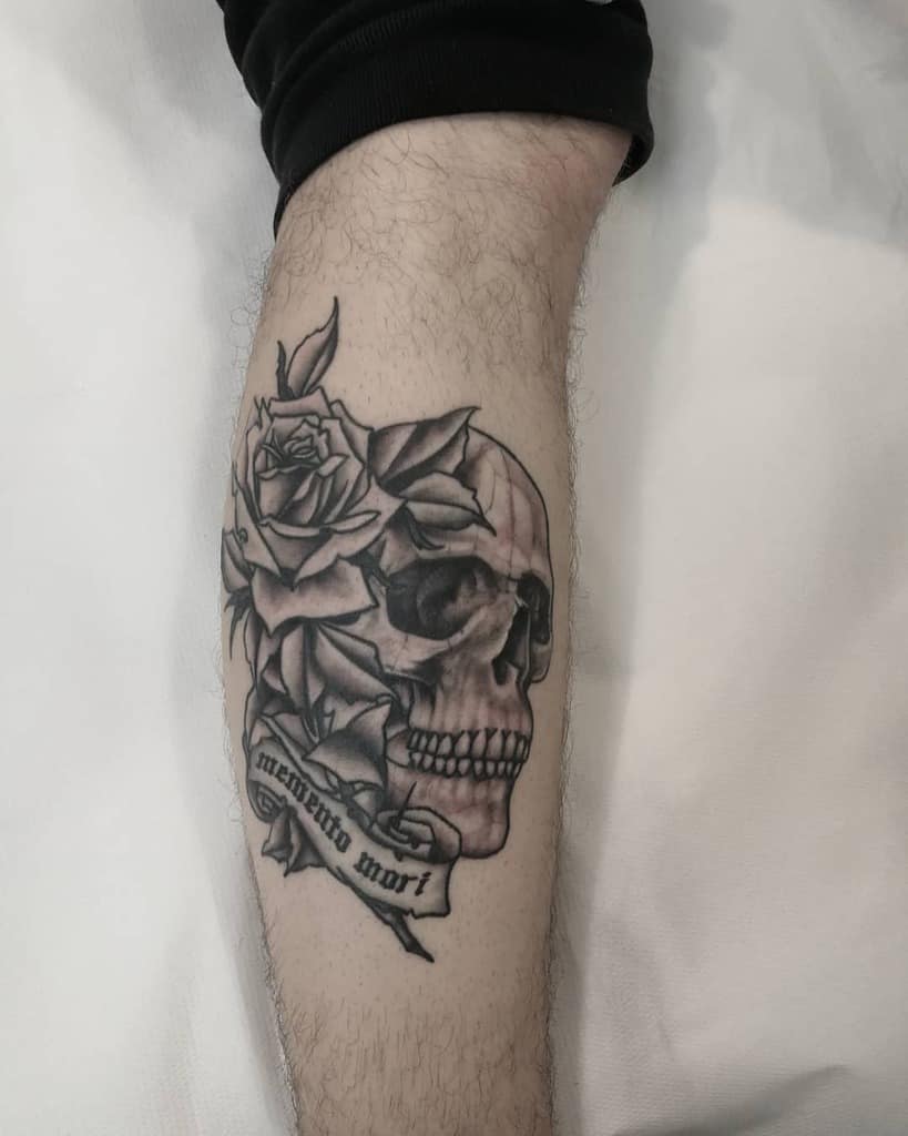 Black and grey tattoo of a skull entwined with roses and a 'Memento Mori' banner, symbolizing mortality and the passage of time.