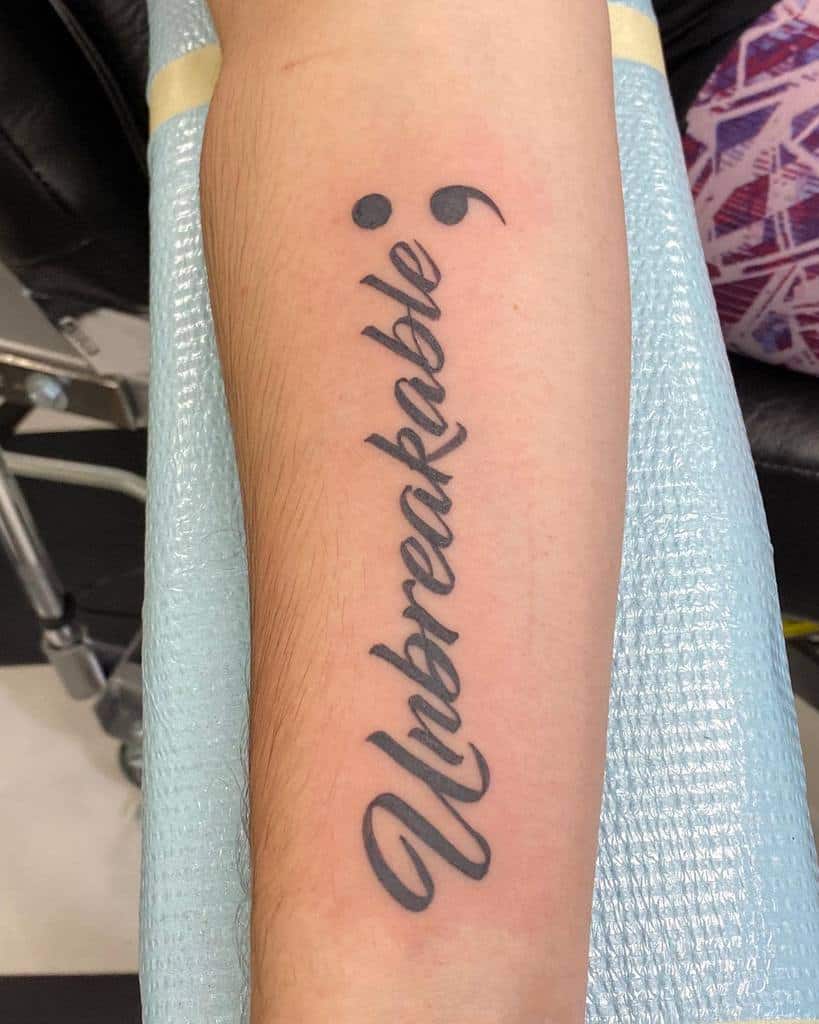 This tattoo features the word "Unbreakable" paired with two semicolons, representing strength, resilience, and overcoming hardships. The semicolon serves as a symbol of continuing life's journey despite challenges.
