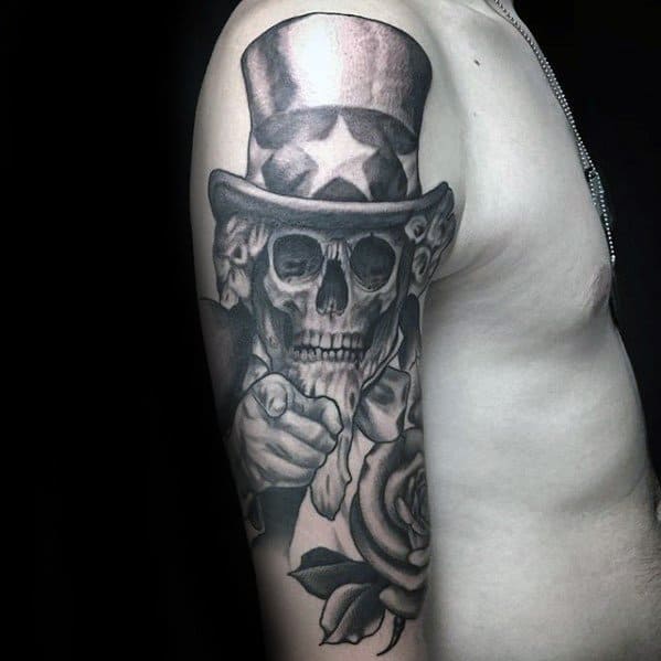 Black and gray Uncle Sam skull tattoo on the upper arm, with bold shading, roses, and a striking pointing pose for a gothic patriotic theme.