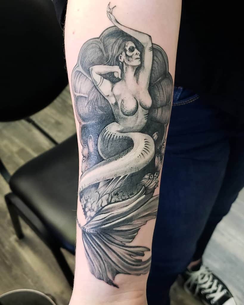 Grayscale tattoo of an undead siren reclining in a seashell, featuring skeletal facial details, flowing hair, and eerie underwater elements