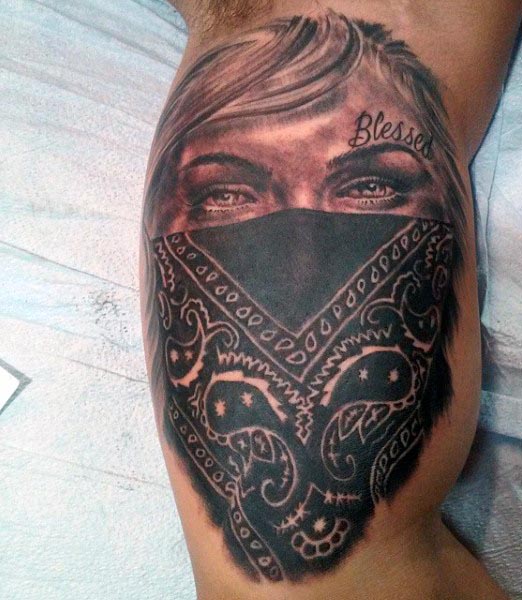 Black and grey tattoo of a woman with intense eyes wearing a paisley-patterned bandana over her face, with the word 'Blessed' above her eyebrow