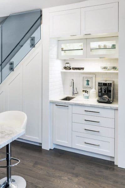 White coffee bar under stairs with built-in cabinets, sink, coffee machine, and illuminated shelves.