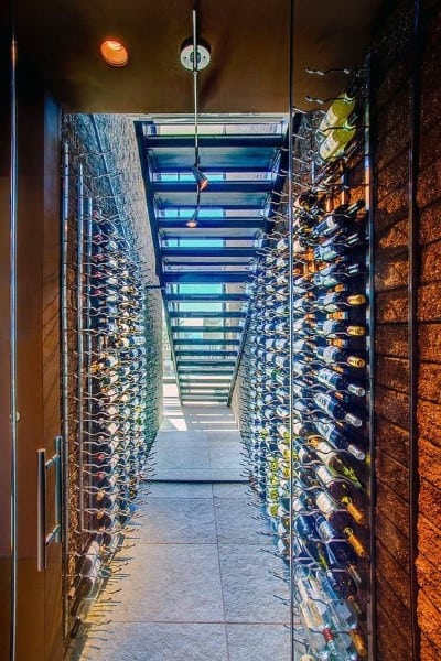A modern wine cellar with wine bottles on wall racks and a glass door entrance, leading to a bright staircase