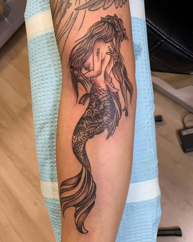 Black and grey tattoo of a mermaid with long flowing hair, intricate scales, and floral details, posed gracefully on the forearm.