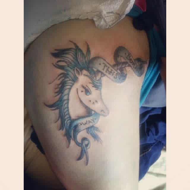Tattoo of a unicorn with flowing mane, surrounded by a 'Born This Way' ribbon, detailed in soft blue and grey tones