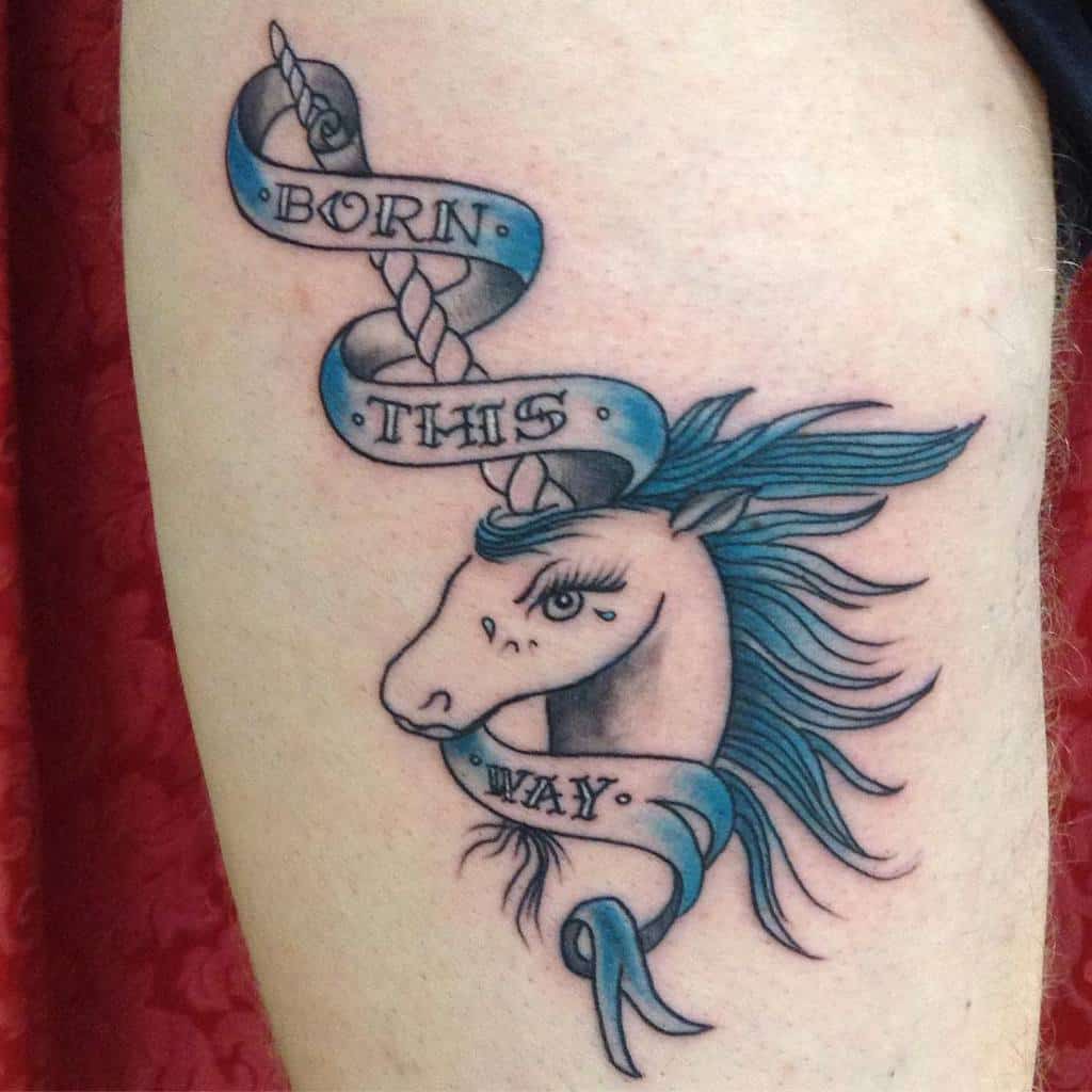 Tattoo of a unicorn head with blue mane, wrapped in a ribbon reading "Born This Way"