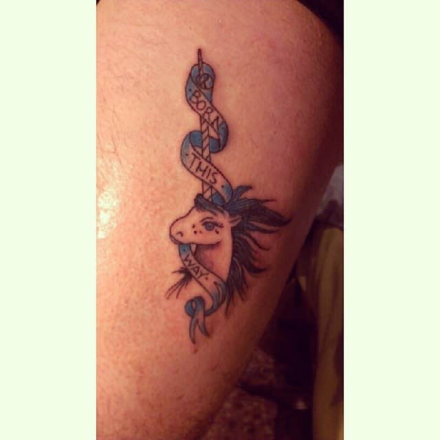 Tattoo of a unicorn with blue mane and a ribbon saying "Born This Way" on a thigh