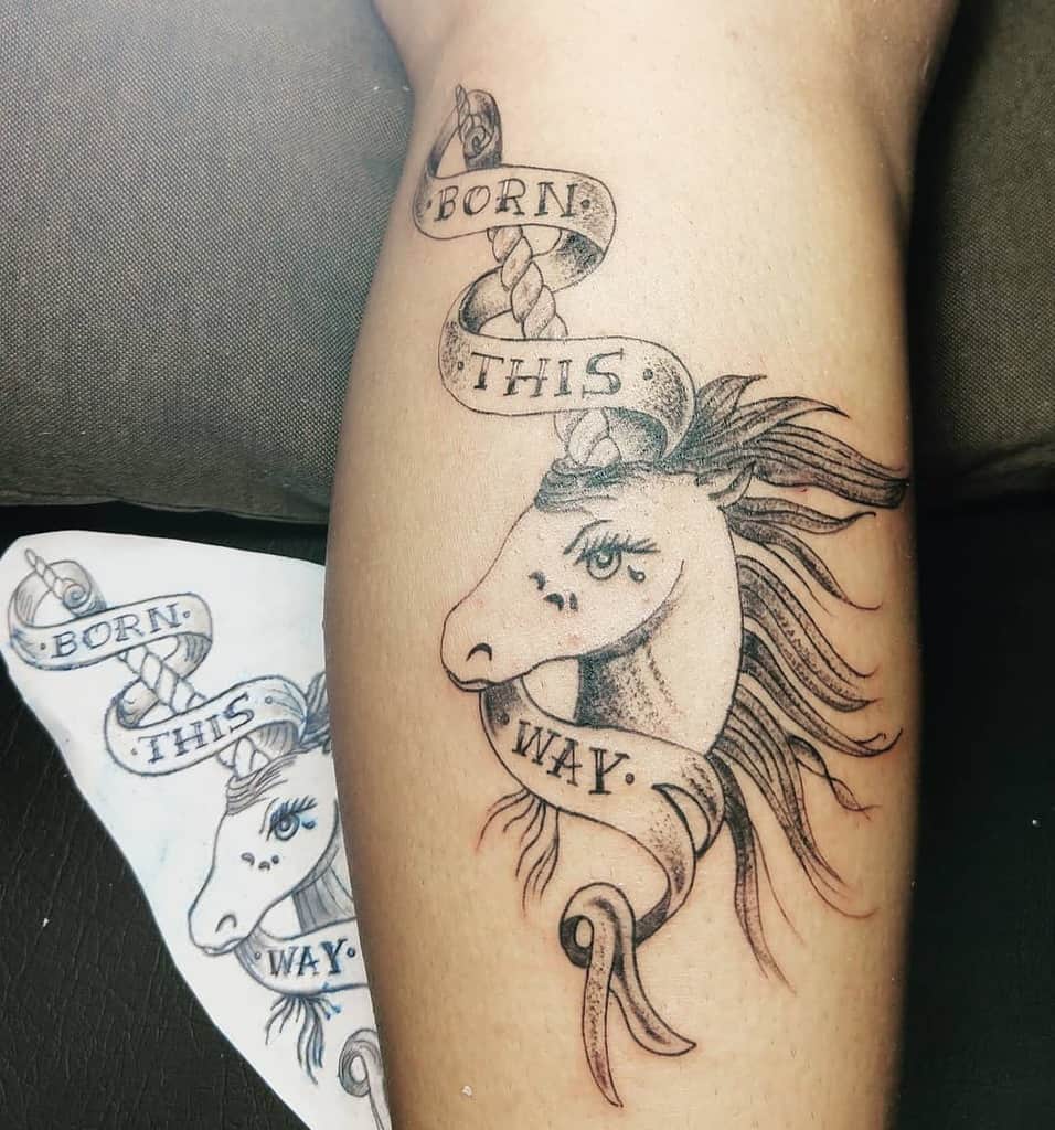 Tattoo of a unicorn head with flowing mane and banner reading "Born This Way" on an arm