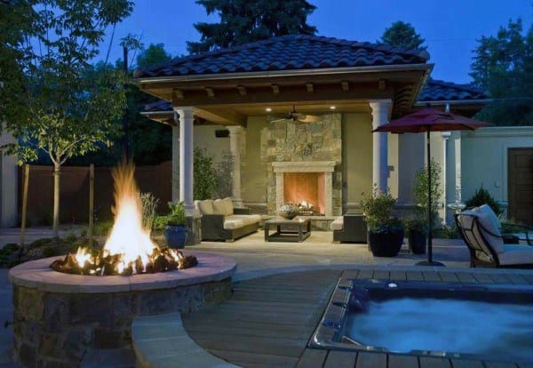 Luxurious pavilion with a stone fireplace, central firepit, cozy seating, and an adjacent hot tub for outdoor leisure.