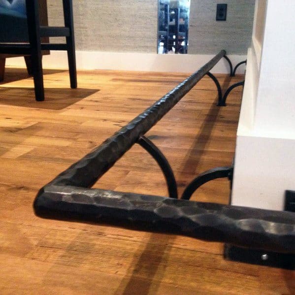Wrought iron foot rail with a hammered texture, mounted on wooden flooring with curved brackets.