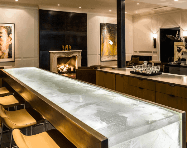 Modern bar with a glowing ice-effect countertop, beige bar stools, and an elegant fireplace.