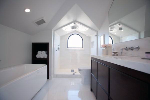 Modern bathroom with vaulted ceiling, freestanding tub, dark vanity, and arched shower window.