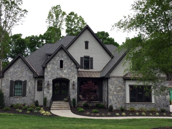 Elegant home with gray stone cladding, gabled rooflines, arched entryway, and a lush front yard featuring a vibrant red tree