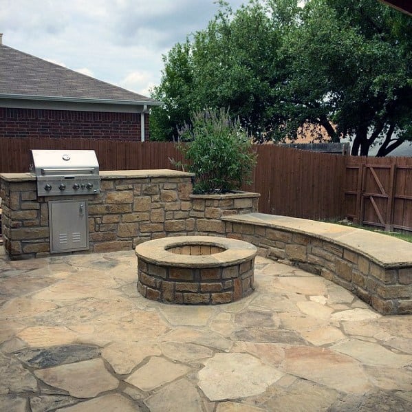 Stone patio with a built-in grill, fire pit, and stone bench, surrounded by a wooden fence and trees