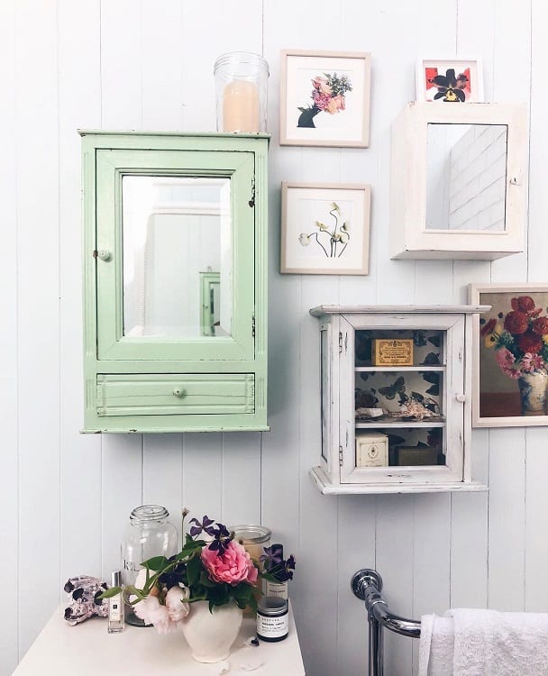 Farmhouse-style vintage bathroom with green and white cabinets, floral art, and decorative items on a white panel wall