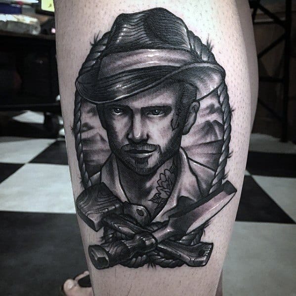 Realistic black and gray tattoo of a rugged man in a fedora, framed by a rope with crossed tools below, symbolizing adventure and craftsmanship.