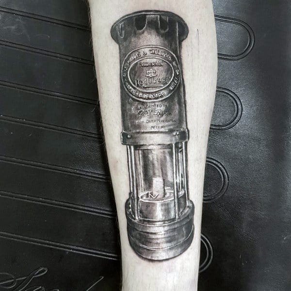 Realistic black and grey tattoo of a vintage Thomas & Williams miner’s lamp with visible serial number and detailed metalwork.