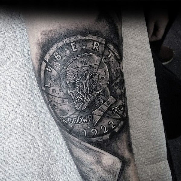 Dark-themed tattoo of a 1922 Liberty coin featuring a cracked skull design, with bold shading for a striking and eerie effect.