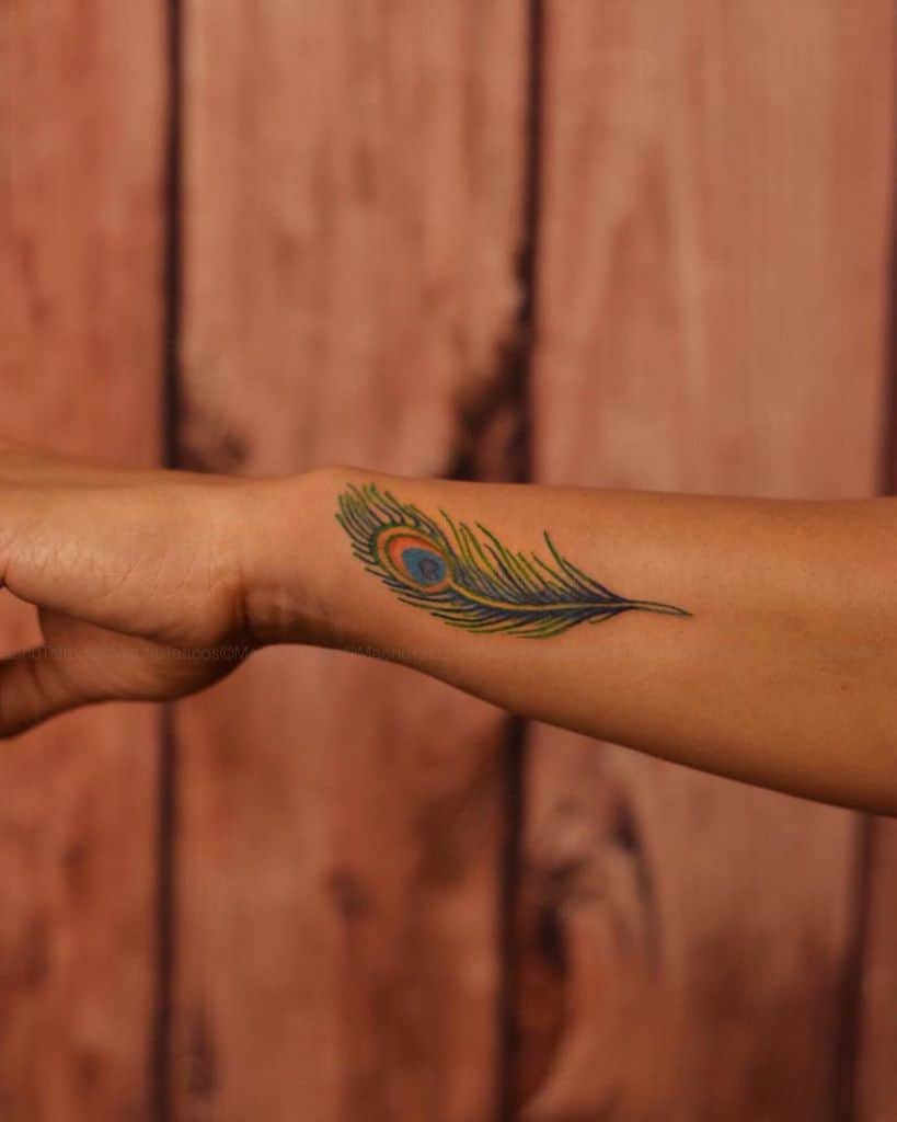 Colorful peacock feather tattoo on the wrist, featuring vibrant green, blue, and orange hues with fine detailing and delicate strokes.