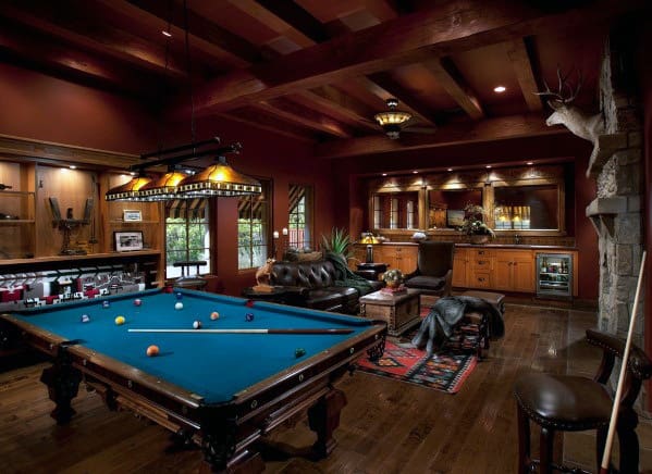 A cozy man cave with a pool table, leather sofa, bar area, and mounted deer head on a wood-paneled wall