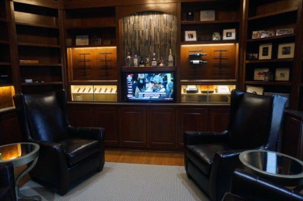Cozy man cave with leather chairs, a TV on, and wooden shelves showcasing small items and collectibles