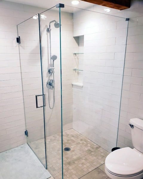 Corner glass shower enclosure with a modern design in a white-tiled bathroom, featuring a showerhead and nearby toilet
