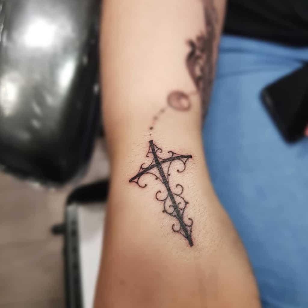 A decorative cross tattoo on the arm featuring intricate scrollwork and fine detailing, creating an ornate and unique design