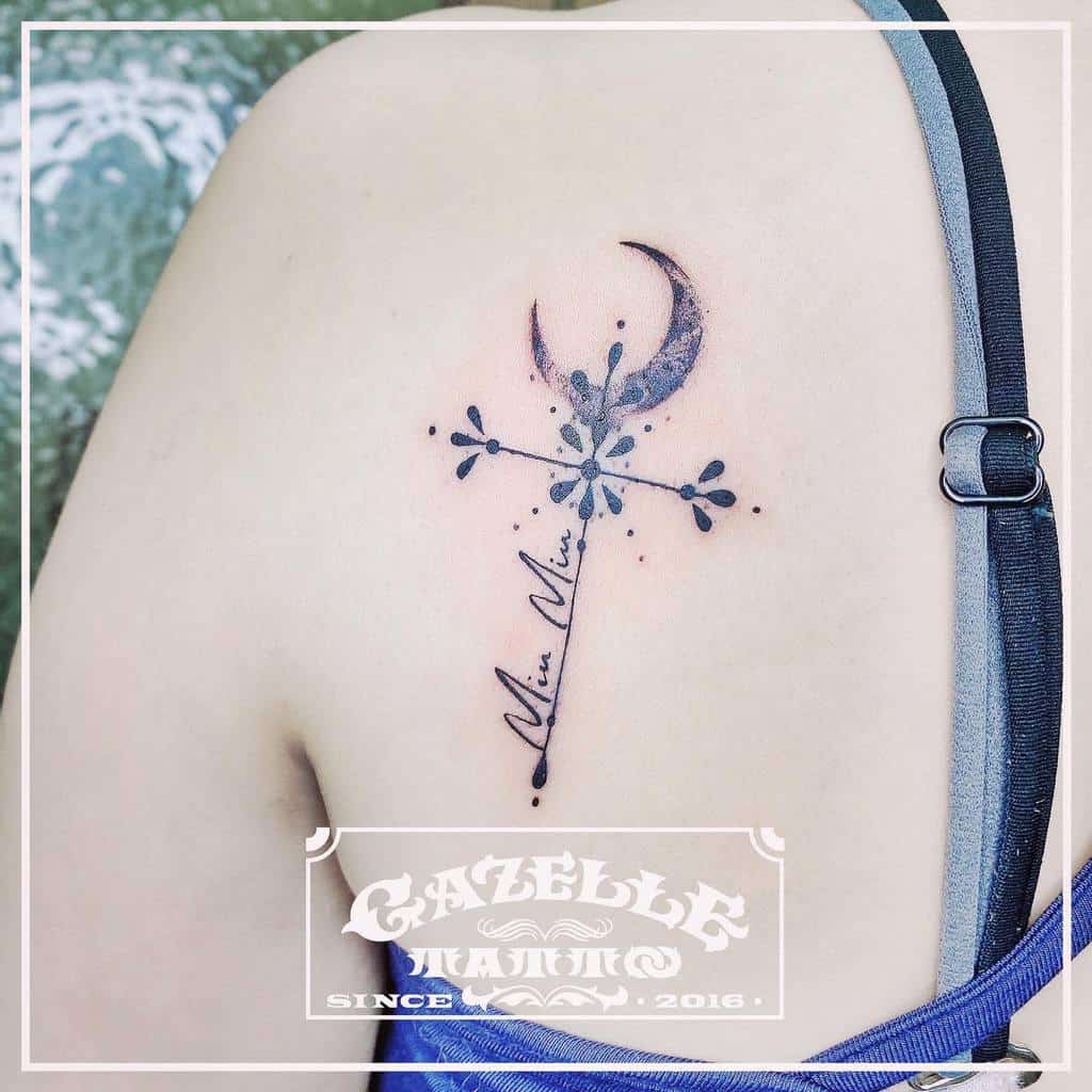 A unique cross tattoo on the upper back featuring floral elements, a crescent moon at the top, and cursive text running vertically along the cross, blending celestial and organic themes
