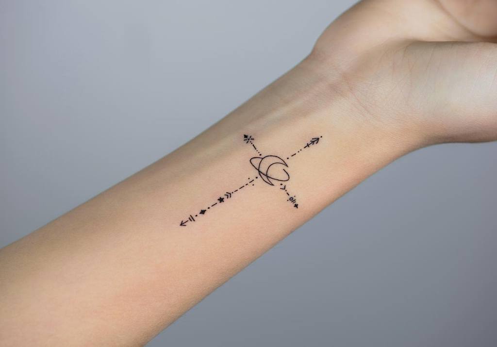 A minimalist tattoo on the wrist featuring a cross formed by arrows and dotted lines, with a crescent moon and planet in the center, blending celestial and geometric elements.