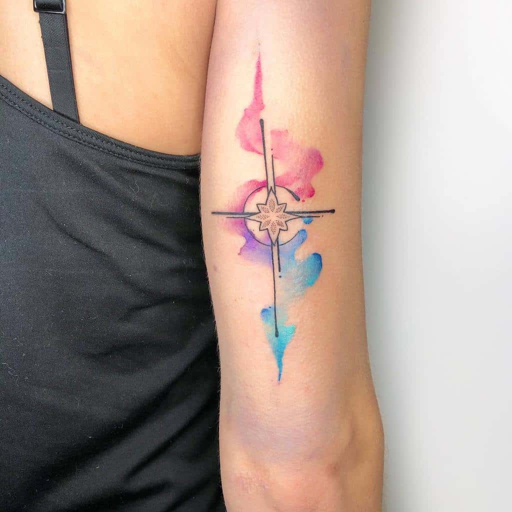 A colorful tattoo on the arm featuring a geometric cross with a central starburst design, accented by watercolor-style splashes in pink, purple, and blue