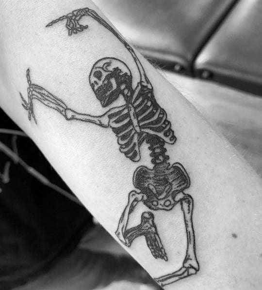 Dancing skeleton tattoo with bold black ink, raised arms, and a playful pose, creating a lively and expressive design on the forearm.