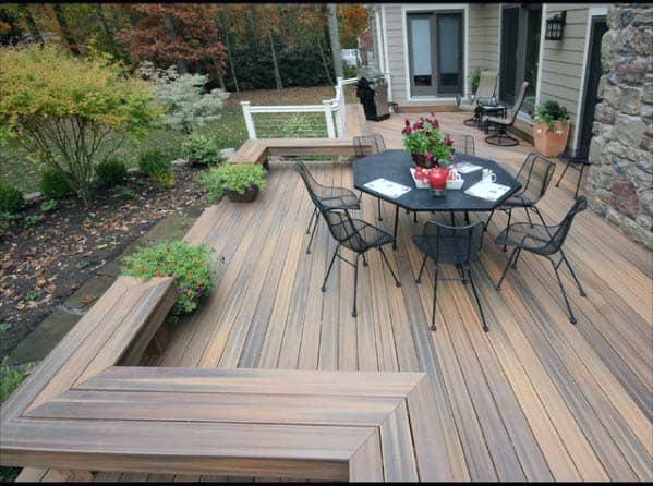 Wooden deck with black table, chairs, and a cozy bench surrounded by lush plants and greenery