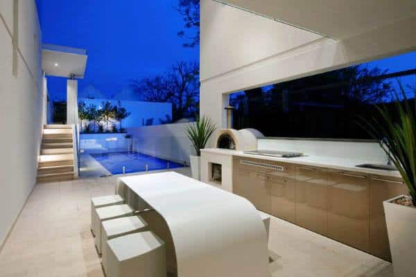 Modern outdoor kitchen with sleek design, dining area, and view of illuminated pool at dusk