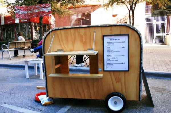 A unique dog house on rolling wheels designed like a food cart, featuring an open window and a menu sign